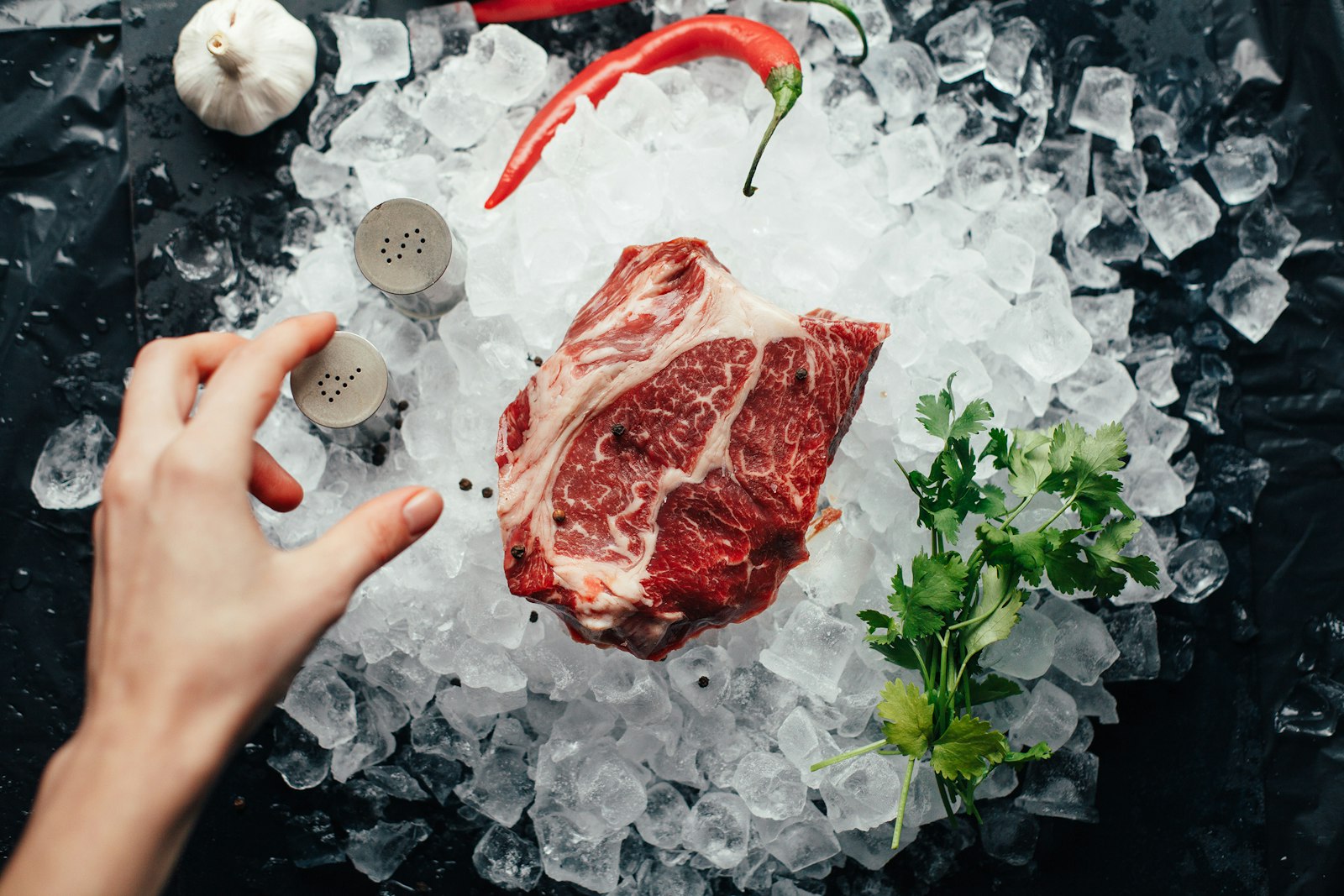 A thick marbled steak resting on crushed ice with a red chile, cilantro and garlic beside it